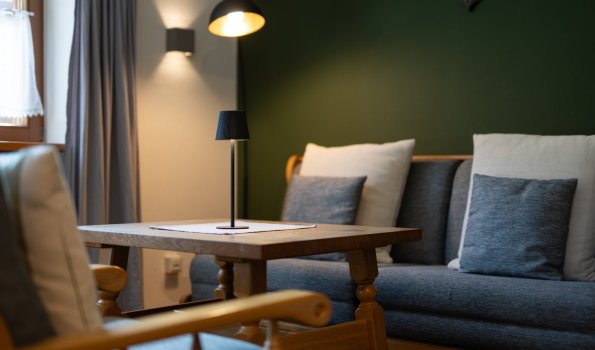 Stilvolle Sitzecke mit Holztisch, Sessel, Sofa mit Kissen und Leuchten vor dunkelgrüner Wand – Bergheimat Ferienwohnungen in Obermaiselstein-Ried im Allgäu.
