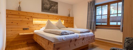 Gemütliches Schlafzimmer mit Doppelbett und Wandverkleidung aus Zirbenholz, Hirschbild und Fenster – Bergheimat Ferienwohnungen in Obermaiselstein-Ried im Allgäu.