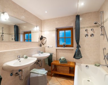 Helles Badezimmer mit Badewanne, Handbrause, zwei Waschbecken, Fenster und Holzhocker – Bergheimat Ferienwohnungen in Obermaiselstein-Ried im Allgäu.