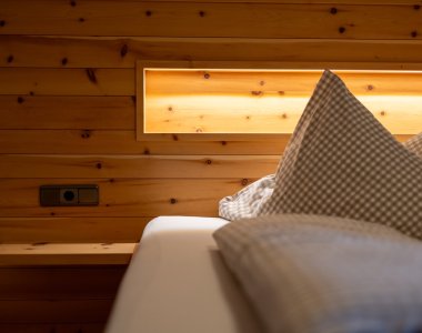 Detail eines Bettes mit Kopfteil aus Zirbenholz, indirekter Beleuchtung und karierten Kissen – Bergheimat Ferienwohnungen in Obermaiselstein-Ried im Allgäu.