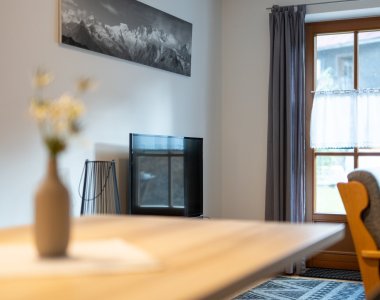 Gemütlicher Wohnbereich mit Holztisch, Blumenvase, Fernseher, Sessel und Bergfoto an der Wand – Bergheimat Ferienwohnungen in Obermaiselstein-Ried im Allgäu.