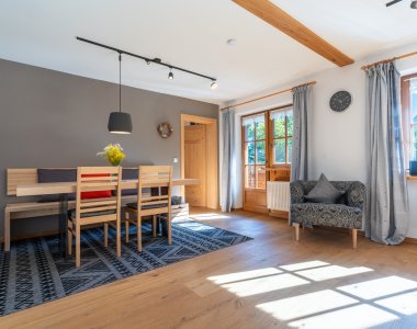 Moderner Essbereich der Bergheimat Ferienwohnungen in Obermaiselstein-Ried mit großem Holztisch, Stühlen, Teppich und Sessel am Fenster.