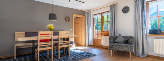 Moderner Essbereich der Bergheimat Ferienwohnungen in Obermaiselstein-Ried mit großem Holztisch, Stühlen, Teppich und Sessel am Fenster.