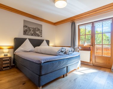 Sonniges Schlafzimmer der Bergheimat Ferienwohnungen in Obermaiselstein-Ried mit Boxspringbett, Nachttischen, Bergbild und Tür zum Balkon.