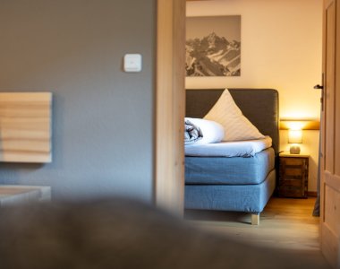Durchblick in ein Schlafzimmer der Bergheimat Ferienwohnungen in Obermaiselstein-Ried mit Boxspringbett, Nachttischlampe und Bergbild an der Wand.