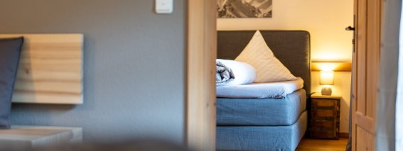 Durchblick in ein Schlafzimmer der Bergheimat Ferienwohnungen in Obermaiselstein-Ried mit Boxspringbett, Nachttischlampe und Bergbild an der Wand.