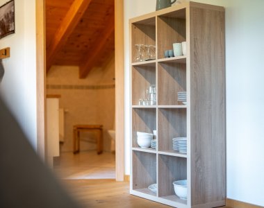 Offenes Regal mit Geschirr und Gläsern in einer Ferienwohnung der Bergheimat Ferienwohnungen in Obermaiselstein-Ried, dahinter der Wohnbereich mit Holzdecke.