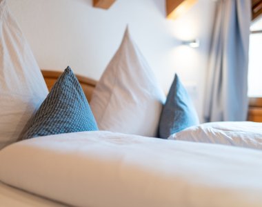 Detailaufnahme eines Doppelbetts in den Bergheimat Ferienwohnungen in Obermaiselstein-Ried: frische weiße Bettwäsche und blau gemusterte Dekokissen.
