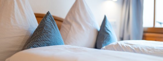 Detailaufnahme eines Doppelbetts in den Bergheimat Ferienwohnungen in Obermaiselstein-Ried: frische weiße Bettwäsche und blau gemusterte Dekokissen.