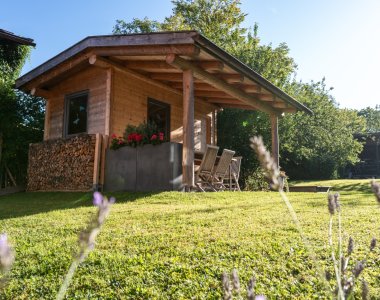 Hölzernes Gartenhaus mit gestapeltem Brennholz, Blumenkasten und Sitzplatz auf der Wiese der Bergheimat Ferienwohnungen in Obermaiselstein-Ried, im Vordergrund blühender Lavendel.