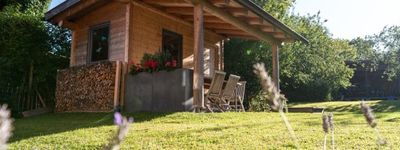 Hölzernes Gartenhaus mit gestapeltem Brennholz, Blumenkasten und Sitzplatz auf der Wiese der Bergheimat Ferienwohnungen in Obermaiselstein-Ried, im Vordergrund blühender Lavendel.