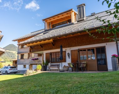Die Bergheimat Ferienwohnungen in Obermaiselstein-Ried an einem sonnigen Tag: Terrasse mit Gartenmöbeln, Rasen und blauer Himmel über dem Allgäu.