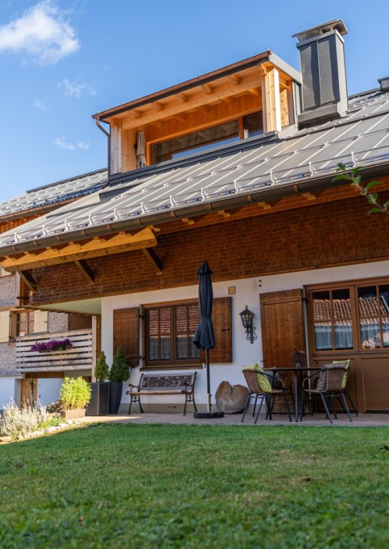 Bergheimat Ferienwohnungen – Haus mit Terrasse bei Sonnenschein Die Bergheimat Ferienwohnungen in Obermaiselstein-Ried an einem sonnigen Tag: Terrasse mit Gartenmöbeln, Rasen und blauer Himmel über dem Allgäu.