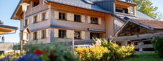 Frontansicht der Bergheimat Ferienwohnungen in Obermaiselstein-Ried: Holzfassade mit Fensterläden und Balkon, im Vordergrund bunte Sommerblumen.