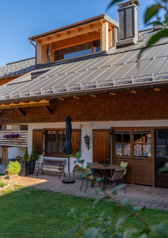 Gartenseite der Bergheimat Ferienwohnungen in Obermaiselstein-Ried mit sonniger Terrasse, Sitzgelegenheiten, Sonnenschirm und gepflegter Rasenfläche vor Bergkulisse.