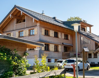 Außenansicht der Bergheimat Ferienwohnungen in Obermaiselstein-Ried: modernes Allgäuer Haus mit Holzschindel-Fassade, Balkonen und Parkplätzen direkt am Haus.