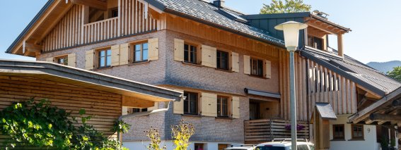 Außenansicht der Bergheimat Ferienwohnungen in Obermaiselstein-Ried: modernes Allgäuer Haus mit Holzschindel-Fassade, Balkonen und Parkplätzen direkt am Haus.
