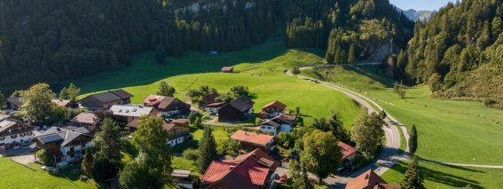 Drohnenaufnahme von Obermaiselstein-Ried im Oberallgäu: Allgäuer Häuser, grüne Almwiesen und bewaldete Berghänge an einem sonnigen Spätsommertag – Lage der Bergheimat Ferienwohnungen.