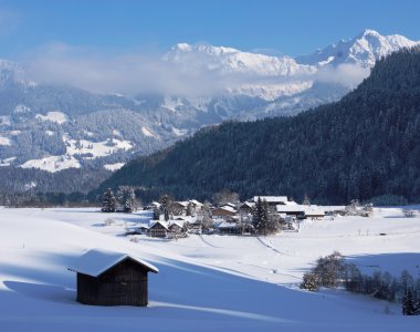 Verschneite Wiesen mit Heustadel und Blick auf Ried und die winterlichen Allgäuer Berge – Bergheimat Ferienwohnungen in Obermaiselstein-Ried im Allgäu.