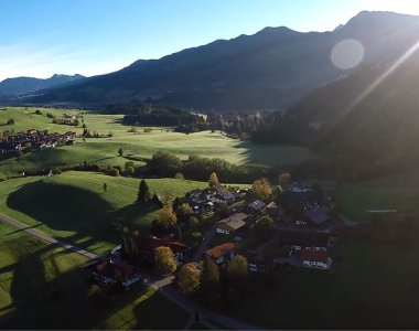 Drohnenaufnahme von Ried bei Obermaiselstein im Gegenlicht mit Wiesen und Bergkette – Bergheimat Ferienwohnungen in Obermaiselstein-Ried im Allgäu.
