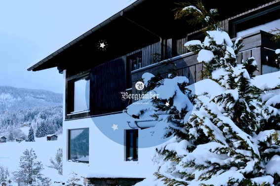 Ein modernes Holzhaus umgeben von schneebedeckten Bäumen in einer ruhigen Winterlandschaft in den Bergen.