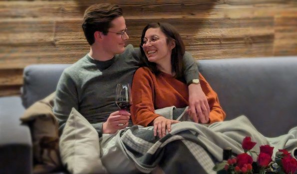 Verliebt auf dem Sofa in einem gemütlichen Raum Ein Paar sitzt auf einem Sofa, genießt ein Glas Wein und lächelt sich an, während sie sich in einer warmen Decke umarmen.