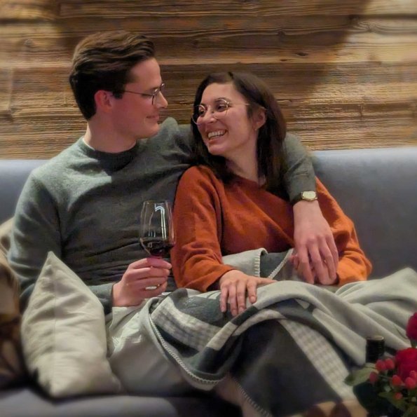 Ein Paar sitzt auf einem Sofa, genießt ein Glas Wein und lächelt sich an, während sie sich in einer warmen Decke umarmen.