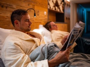 Zwei Personen entspannen in einem Hotelzimmer und lesen Zeitungen. Es herrscht eine ruhige Atmosphäre und angenehme Beleuchtung.
