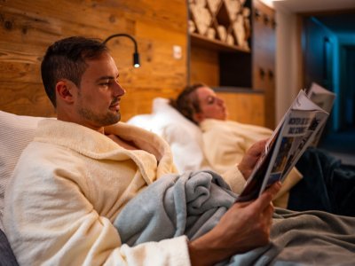 Zwei Personen entspannen in einem Hotelzimmer und lesen Zeitungen. Es herrscht eine ruhige Atmosphäre und angenehme Beleuchtung.