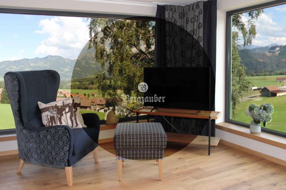 In einem Raum steht ein Stuhl und ein Tisch. Draußen sind Berge und Bäume zu sehen. Es ist tagsüber und hell.