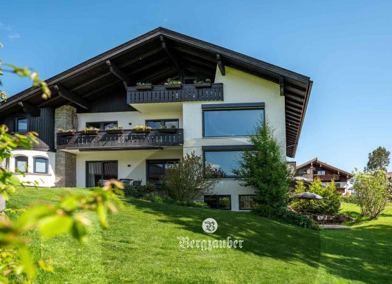 Ein modernes Haus steht in einer grünen Umgebung mit Bäumen. Die Sonne scheint klar auf die Fassade.
