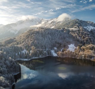 Eine verschneite Landschaft um einen stillen See. Die Sonne strahlt über die Berge, während Wolken den Himmel zieren.