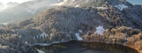 Eine verschneite Landschaft um einen stillen See. Die Sonne strahlt über die Berge, während Wolken den Himmel zieren.