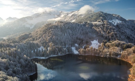 Eine verschneite Landschaft um einen stillen See. Die Sonne strahlt über die Berge, während Wolken den Himmel zieren.