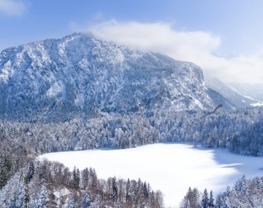 Eine ruhige Winterlandschaft zeigt einen gefrorenen See, umgeben von schneebedeckten Bäumen und majestätischen Bergen.