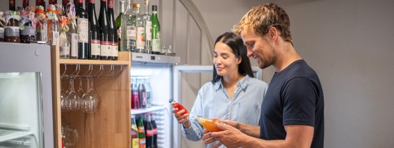 Ein Paar steht im Vorratsraum und wählt Getränke aus dem Kühlschrank. Sie schauen auf ihr Handy.
