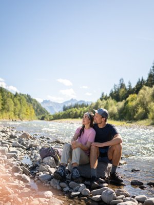 Ein Paar sitzt am Fluss und schaut auf die Berge. Die Sonne scheint und die Landschaft ist grün.