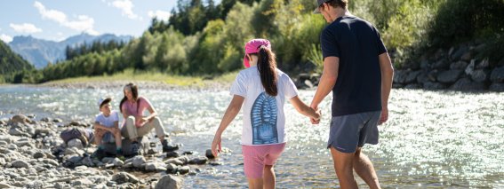 Eine Familie steht am Fluss und geht über Steine. Kinder spielen im Wasser und Erwachsene beobachten sie dabei.