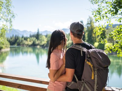 Ein Paar steht an einem Holzgeländer am See. Sie schauen auf das Wasser und in die Berge. Es ist ein sonniger Tag.