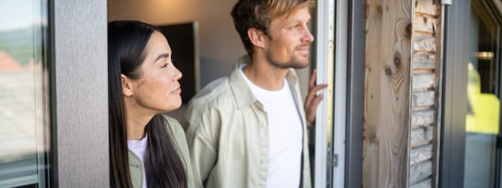 Ein Paar steht am Fenster und schaut nach draußen. Es ist Tag und die Umgebung ist grüner.