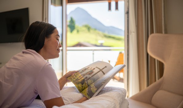 Eine Frau sitzt im Zimmer und liest eine Zeitschrift. Sie schaut in Richtung Fenster.