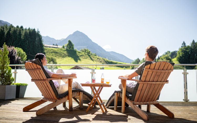 Zwei Personen sitzen auf einer Terrasse und genießen die Aussicht auf die Berge. Es ist ein sonniger Tag.