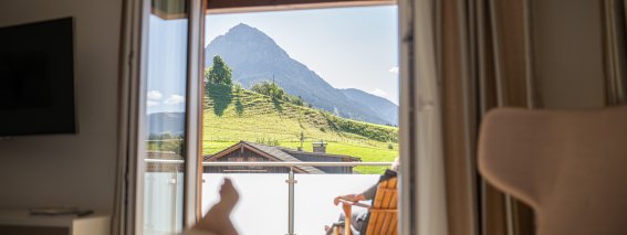 Die Sonne scheint durch das Fenster eines Zimmers. Eine Person sitzt draußen und schaut auf die Berge und die grüne Landschaft.