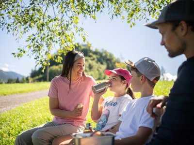 Eine Familie sitzt auf einer Wiese und trinkt. Es ist ein sonniger Tag mit Bäumen im Hintergrund.