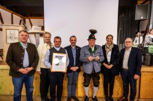 Sechs Personen stehen auf der Bühne, sie halten eine Auszeichnung und einen Pokal in der Hand. Es ist Abend und eine Feier.