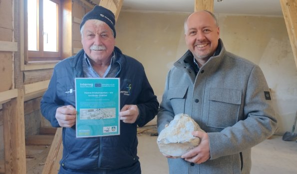 Unterzeichnung des Partnerschaftsvertrags für die Klamm-Erlebniswelten Zwei Männer halten einen Stein und ein Informationsblatt in einem renovierten Raum. Der Ort hat Holzrahmenwände und ist hell.