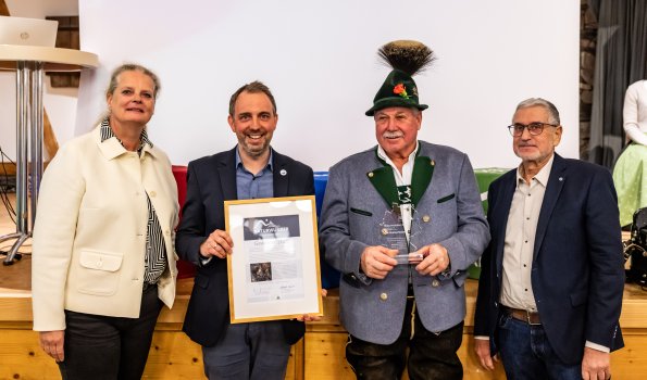 Übergabe Naturwunderwahl 2025 Vier Personen stehen auf einer Bühne und präsentieren eine Urkunde. Es ist eine feierliche Veranstaltung in einem Saal.