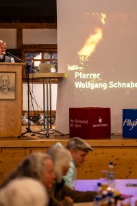 Pfarrer spricht vor einer Gruppe Menschen in einem Saal. Die Veranstaltung findet am Abend statt.