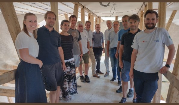 Info-Workshop Teilnehmer Eine Gruppe von Menschen steht für ein Foto in einem Halbkreis inmitten einer Baustelle. Im Hintergrund erkennt man Holzpflöcke.
