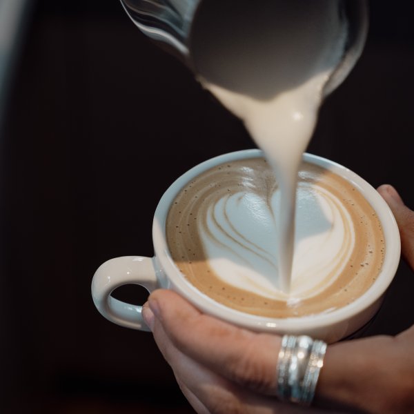 Cappuccino wird in eine Tasse gegossen Eine Person gießt heiße Milch in einen Kaffee und formt ein Herz auf der Oberfläche. Der Moment zeigt Handarbeit in einem Café.